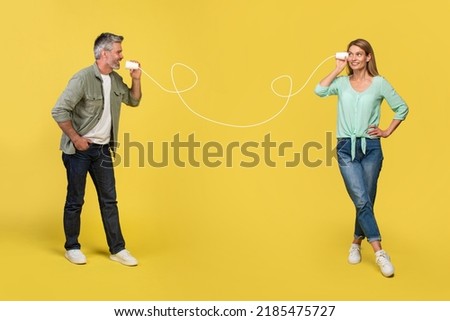 Caucasian middle aged man telling something to his wife through tin can phone with painted string, standing over yellow background, creative collage Royalty-Free Stock Photo #2185475727