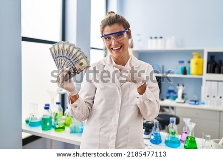 Young woman working at scientist laboratory holding money smiling happy pointing with hand and finger 