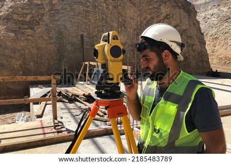 Land surveyor stands by theodolite on a tripod against construction site at background. Topographer does measurements on geodetic total station. Geodetic or topographic works Royalty-Free Stock Photo #2185483789