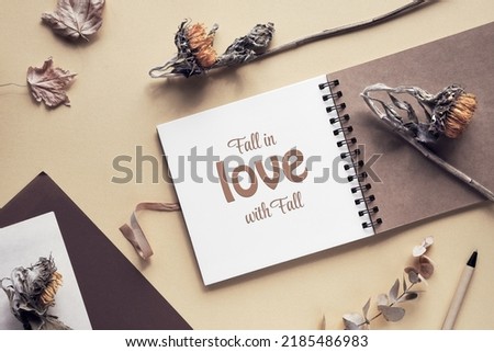 Caption motivator text Fall in love with Fall in square album, notebook with spiral binder. Wooden pen, dry sunflower flowers and maple leaves. Soft natural light. Monochromatic beige color hues.