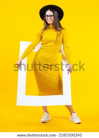 Young woman. Girl with white frame. Model brunette in full growth. Female student in dress and sneakers. Woman posing on yellow. Business lady in long dress and hat. Fashion lady peeking out frame
