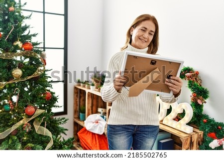Middle age caucasian woman looking picture standing by christmas tree at home