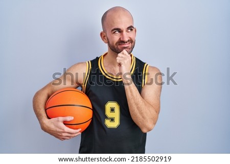 Young bald man with beard wearing basketball uniform holding ball with hand on chin thinking about question, pensive expression. smiling and thoughtful face. doubt concept. 