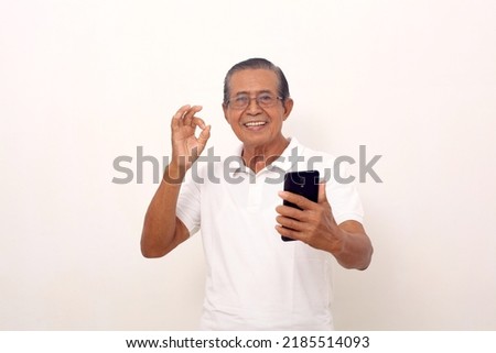 Happy asian old man standing while holding a cell phone and showing okay hand gesture