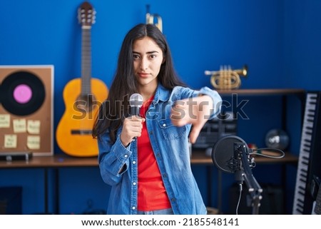 Young teenager girl singing song using microphone with angry face, negative sign showing dislike with thumbs down, rejection concept 
