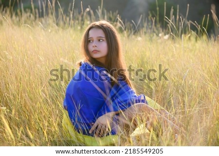 A teenage girl sits in tall wild grass on the slope of a ravine with a Ukrainian flag on her shoulders.