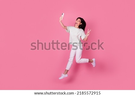 Photo of cute young japanese lady hold gadget take selfie jump show v-sign dressed stylish white clothes isolated on pink color background