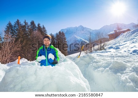 Happy young boy in ski winter outfit prepare snowballs in fortress with high mountains on background