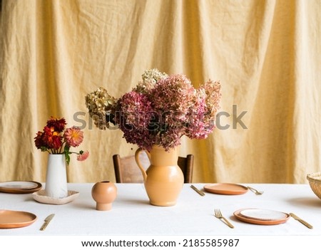 
 A bouquet of dry hydrangea flowers stands in a vase in the middle of the table against a yellow fabric background. Ceramic dishes and cutlery are placed on the table. Horizontal photo