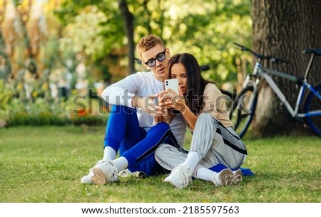 Young modern couple is using mobile technology when sitting in the park. A female is taking photos on her smartphone, a man is looking at the phone and talking to a woman. Bikes on the background.