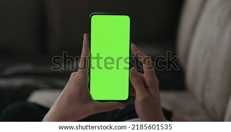 Young woman lying on a couch and using smartphone with vertical green screen, wide photo