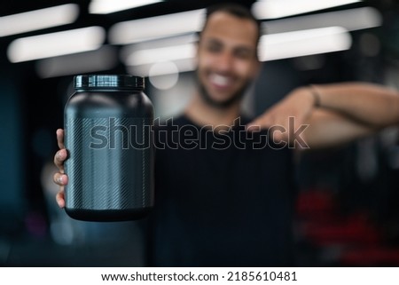 Black Sporty Male Demonstrating Blank Container For Whey Protein Powder And Pointing At It While Standing At Gym, Smiling African American Sportsman Recommending Fitness Supplements, Selective Focus Royalty-Free Stock Photo #2185610481