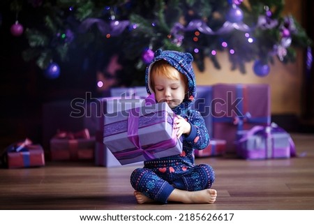 A cute girl in a purple jumpsuit opens a box with a Christmas present while sitting on the floor near the Christmas tree in a cozy living room on Christmas Eve.