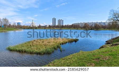 There are trees and grass on the shore of the lake, part of the shore is concreted. There is a small island in the water. On the opposite shore is an Orthodox church and residential houses. Sunny Royalty-Free Stock Photo #2185629057
