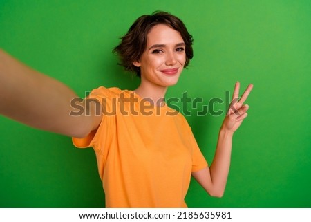 Photo of dreamy funny young woman dressed orange t-shirt showing v-sign tacking selfie isolated green color background