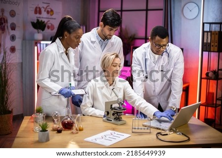 Team of medical research scientists work on a new generation disease cure. They use microscope, test tubes, laptop and writing down analysis results. Laboratory looks busy, modern, night time.