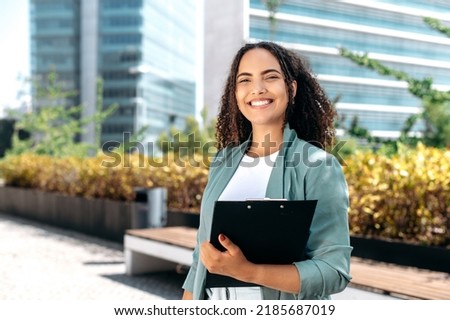 Elegant confident young woman with curly hair, formally dressed, stands outdoors against the background of the business center, holding a folder with documents, looks at camera, smiles Royalty-Free Stock Photo #2185687019