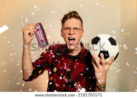 Ecstatic fan, young man holding ticket for soccer match and soccer ball. Confetti in the back. Man winning ticket as prize  Royalty-Free Stock Photo #2185705571