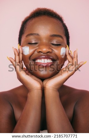 Portrait of smiling black woman keeping her eyes closed applying face cream onto her cheeks isolated over pink background. Beauty woman face skin care. Facial