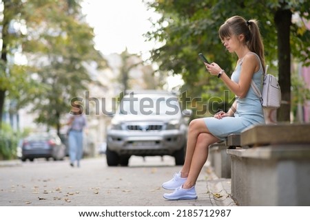 Young pretty woman messaging on mobile phone on warm summer day sitting on a city street bench outdoors.