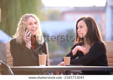 Happy young woman chatting on mobile phone ignoring her bored friend. Female friends sitting at street cafe outdoors and having hard time communicating with each other. Friendship problems concept Royalty-Free Stock Photo #2185713045