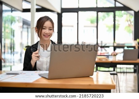 Attractive young Asian businesswoman or female consultant at her office desk sipping morning coffee while using laptop computer.