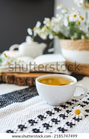 Cup of chamomile tea, open book, wicker basket with flowers, atmospheric photo Royalty-Free Stock Photo #2185747499
