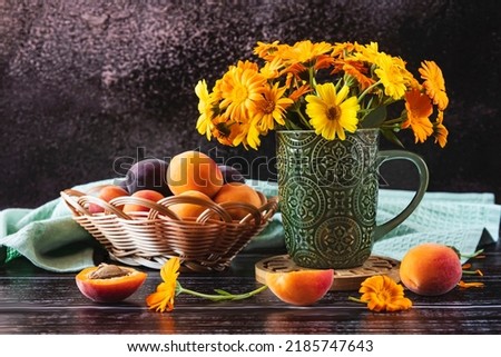 Yellow calendula flowers in a green cup and apricots in a basket on a dark background. Side view.