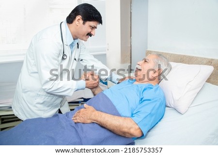 Smiling Indian caring doctor supporting putting hand on shoulder of olde senior male patient lying on bed at clinic or hospital. Elderly people health care concept.
