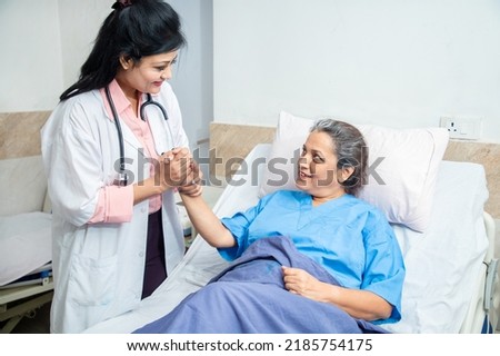 Smiling Indian caring doctor supporting holding hand of olde senior female patient lying on bed at clinic or hospital. Elderly people health care concept. Royalty-Free Stock Photo #2185754175