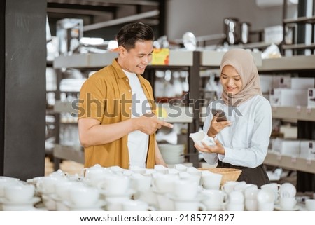 Young woman in headscarf using smartphone camera to photograph cups with her male friends in glassware shop