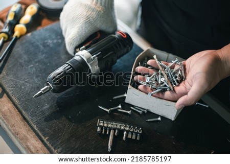 Close up asian man hand holding electric cordless screwdriver machine and screws lie for screwing a screw assembling furniture at home. iron screw, screws as a background, wood screw, concept industry Royalty-Free Stock Photo #2185785197