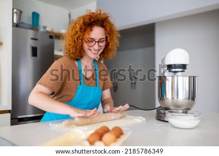 Happy attractive young adult woman housewife baker wear apron holding pin rolling dough on kitchen table baking pastry concept cooking cake biscuit doing bakery making homemade pizza at home