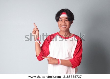 Portrait of attractive Asian man in t-shirt with red and white ribbon on head, pointing at something with finger. Isolated image on gray background