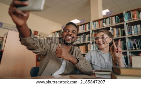 African merican guy and caucasian girl have fun smiling and taking selfie photos on smartphone camera at university library indoors. Cheerful students have rest while prepare for examination