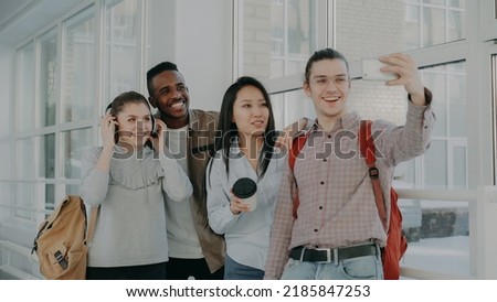 Group of four multi-ethnic students taking selfie on smartphone camera while standing in corridor of university . Hipster guy holding phone and friends are posing positively