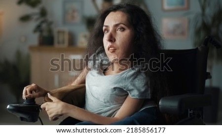 Frustrated woman with spinal muscular atrophy sitting in a wheelchair in a dim living room illuminated by a lamp Royalty-Free Stock Photo #2185856177