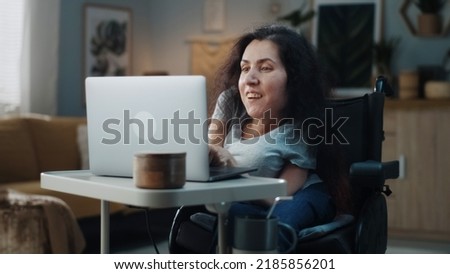Happy woman in a wheelchair sitting at the table and editing video for her blog on a laptop at home studio, for filming near the tripod with ring LED lamp