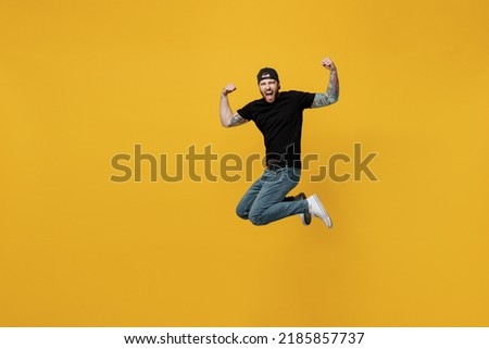 Full body young overjoyed cool bearded tattooed man 20s he wears casual black t-shirt cap jump high do winer gesture isolated on plain yellow wall background studio portrait. People lifestyle concept Royalty-Free Stock Photo #2185857737