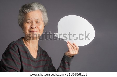 Speech bubble concept. An elderly Asian woman in a sweater with short gray hair holding a blank speech bubble and looks at the camera with a smile while standing on a gray background