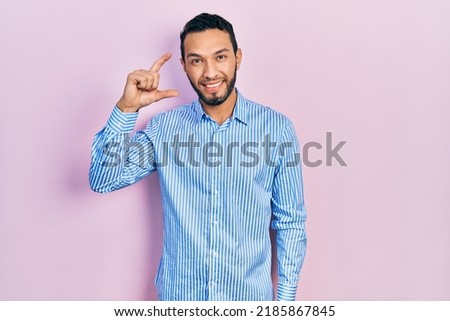 Hispanic man with beard wearing casual blue shirt smiling and confident gesturing with hand doing small size sign with fingers looking and the camera. measure concept. 