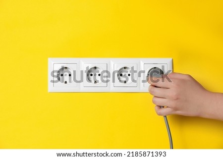 Woman inserting plug into power socket on yellow wall, closeup. Electrical supply Royalty-Free Stock Photo #2185871393