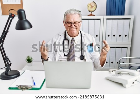 Senior caucasian man wearing doctor uniform and stethoscope at the clinic gesturing finger crossed smiling with hope and eyes closed. luck and superstitious concept. 