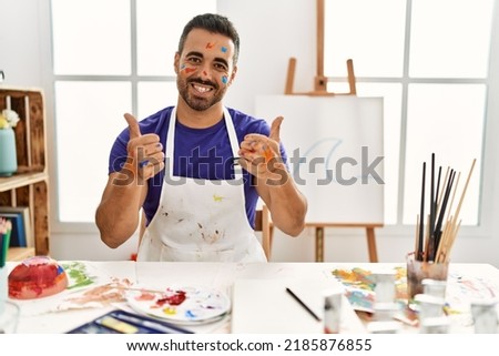 Young hispanic man with beard at art studio with painted face success sign doing positive gesture with hand, thumbs up smiling and happy. cheerful expression and winner gesture. 