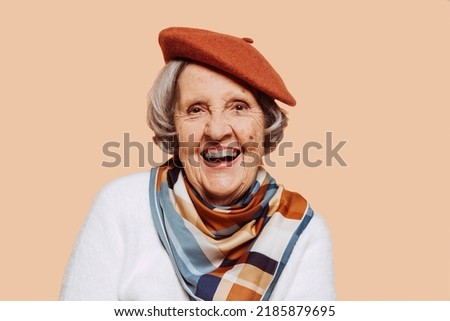 Happy smiling elderly fashionable grandmother wears brown beret, white sweater and colorful scarf, studio portrait. Senior old retired woman looking at camera over beige background.