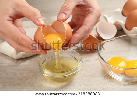 Woman separating egg yolk from white over glass bowl at wooden table, closeup Royalty-Free Stock Photo #2185880631