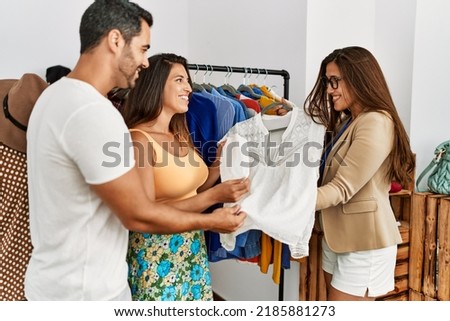 Young hispanic shopkeeper woman showing shirt to hispanic couple at clothes store.
