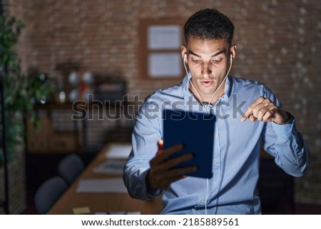Handsome hispanic man working at the office at night pointing down with fingers showing advertisement, surprised face and open mouth 
