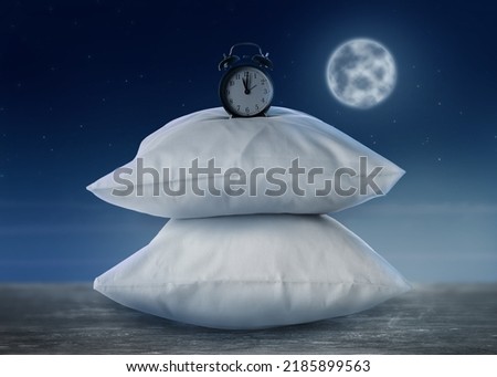Soft pillows and alarm clock on grey table against night sky with full moon. Insomnia Royalty-Free Stock Photo #2185899563