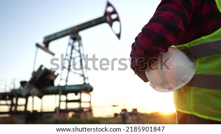 oil production. a worker holding a protective hard hat at sunset in the background an oil pump. oilfield business a extraction concept lifestyle. oil extraction pump. oil pump rig Royalty-Free Stock Photo #2185901847